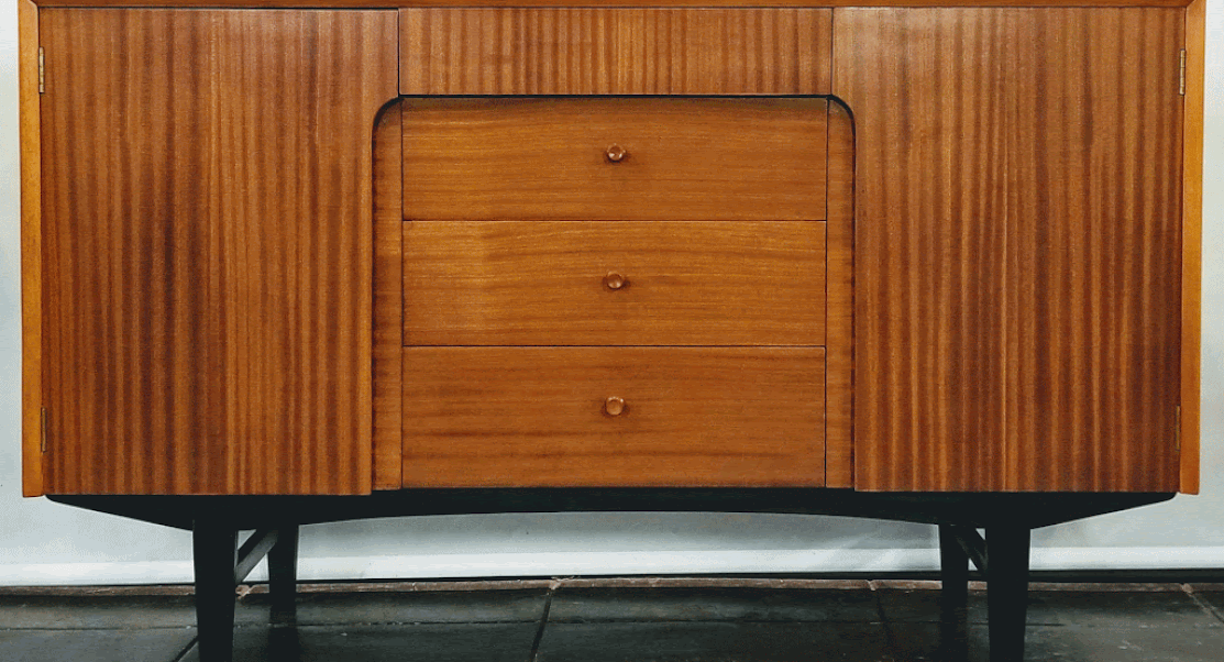 Mid-Century UK Modern credenza by The Co-Operative Wholesale Society has shelved single-door cabinets on each side and four drawers at center.  The oak carcase is veneered with ribbon cut Sapele wood from Africa. The trim and knobs are Birch wood.