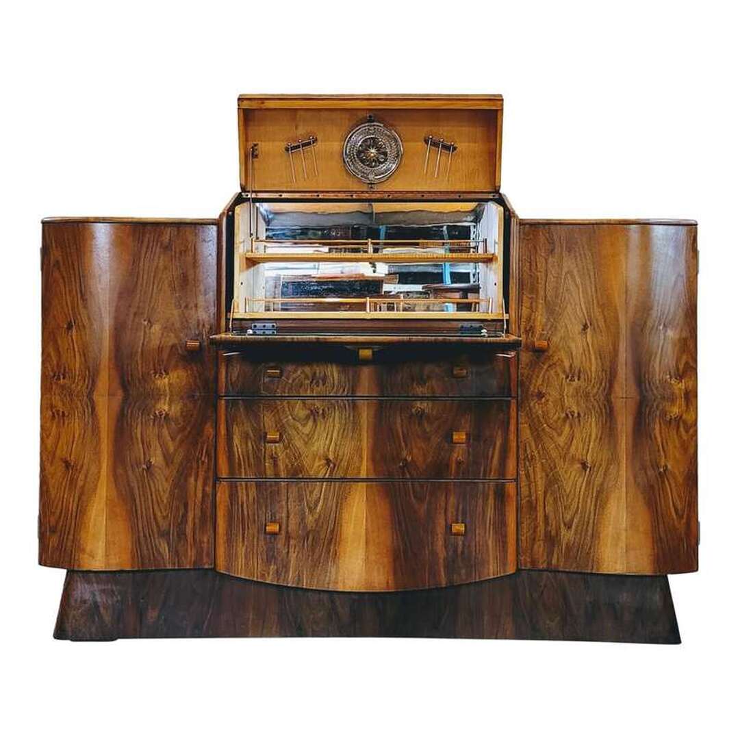 English walnut-finished cocktail sideboard from the 1950s blends Classic and Art Deco styling for an updated Traditional Mid-Century design.  The stepped top contains a central cocktail cabinet that opens with a tug on the brass and Catalin pull on the front.  The interior of the cocktail cabinet has a mirrored back and rear of fall-front door.  Two storage shelves for drinking glasses, etc. feature copper galleries.  The drinks prep station hardware is all copper, including the piano hinge at top, interior drop hinges, galleries, and cocktail pick caddies. Six new stainless steel cocktail picks are housed in the copper caddies under the top of the cabinet along with a stainless steel citrus reamer.  Three storage drawers are fitted below the cocktail cabinet. The top drawer is fitted with spacers for organizing. The two lower drawers are nice and deep, offering plenty of storage.  On each side of the cocktail cabinet and drawers are curved-front single-door storage cabinets. Each cabinet is fitted with one standard shelf.  The carcase is supported by a graceful curved and splayed plinth base.  The carcase is constructed from oak wood.  The cabinet door interiors and curved base trim are mahogany wood.  The interior of the cocktail cabinet is maple wood.  The piece is finished with matched burl figured English walnut wood veneers.  The marbleized butterscotch cylindrical knobs are Catalin thermoplastic. The knob on the front of the cocktail cabinet is placed over an elliptical brass escutcheon.
